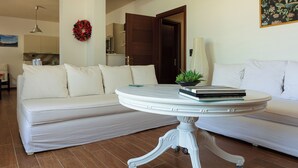 A living room with a white sofa, a round table with books, a potted plant, and a door leading to another room.