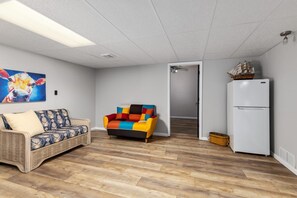 Una habitación con un sofá de mimbre, un sillón colorido, un refrigerador blanco y un modelo de barco encima.