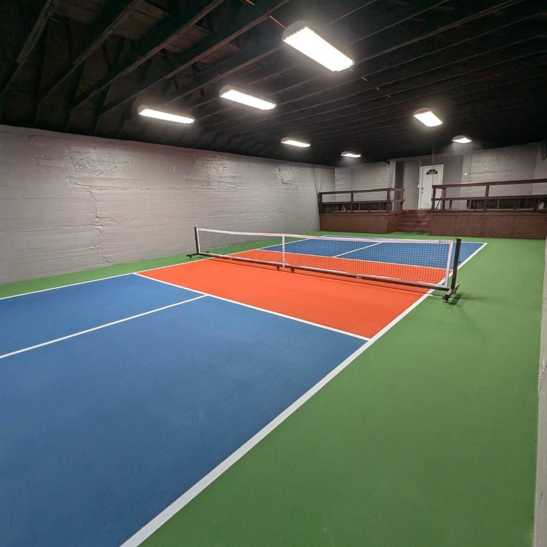 Un court de tennis intérieur doté d’un filet, d’une surface de jeu verte et bleue, et d’un éclairage au plafond.