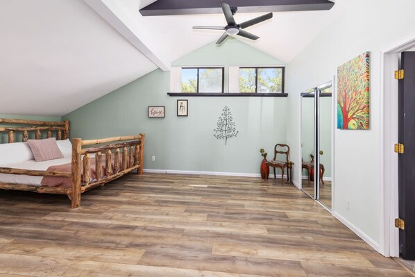Una habitación con techo inclinado, cama con cabecera de madera, ventilador de techo y un ventanal grande.
