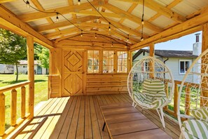 A wooden porch with a covered roof, a hanging chair, and a view of a house in the background.