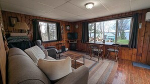 A cozy cabin interior with a sofa, TV, dining table, and large windows. - 1BR Cozy Cabin- Small walk from Chippewa Lake (Chippewa Lake)