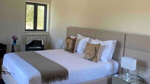 A neatly made bed with white linens and decorative pillows, a bedside table with a lamp, and a window with a view of greenery. - Lisbon country house  (Alcochete)