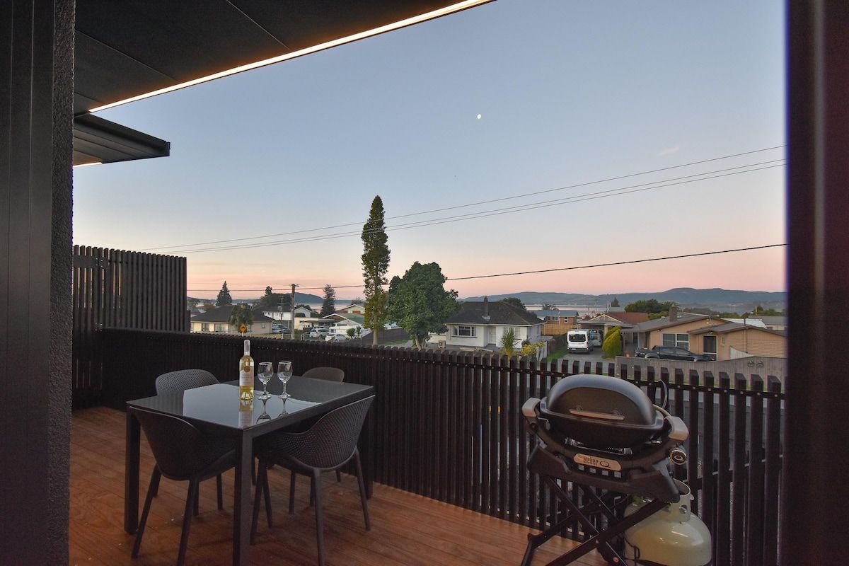A balcony with a table and chairs, a grill, and a view of houses and a tree.
