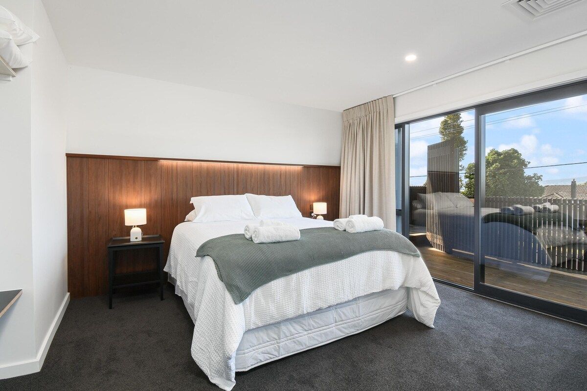 A modern hotel room with a large bed, a bedside table with a lamp, and a view through a large window.