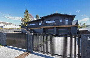 A modern two-story building with a flat roof, a balcony on the upper floor, and a gated entrance. - The Bluebird - Lake Views, Stylish, Sleeps 4 (Rotorua)