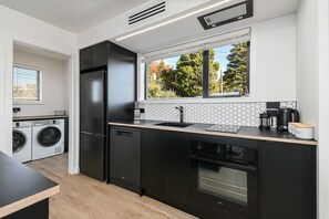 A modern kitchen with a built-in oven, a dishwasher, and a large window with a view of trees. - The Bluebird - Lake Views, Stylish, Sleeps 4 (Rotorua)