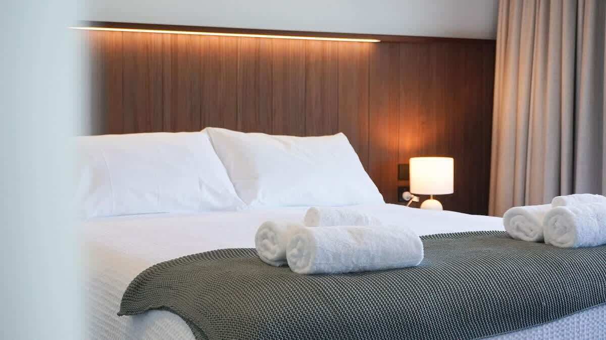 A hotel room with a large bed, a bedside lamp, and rolled towels on the bed.