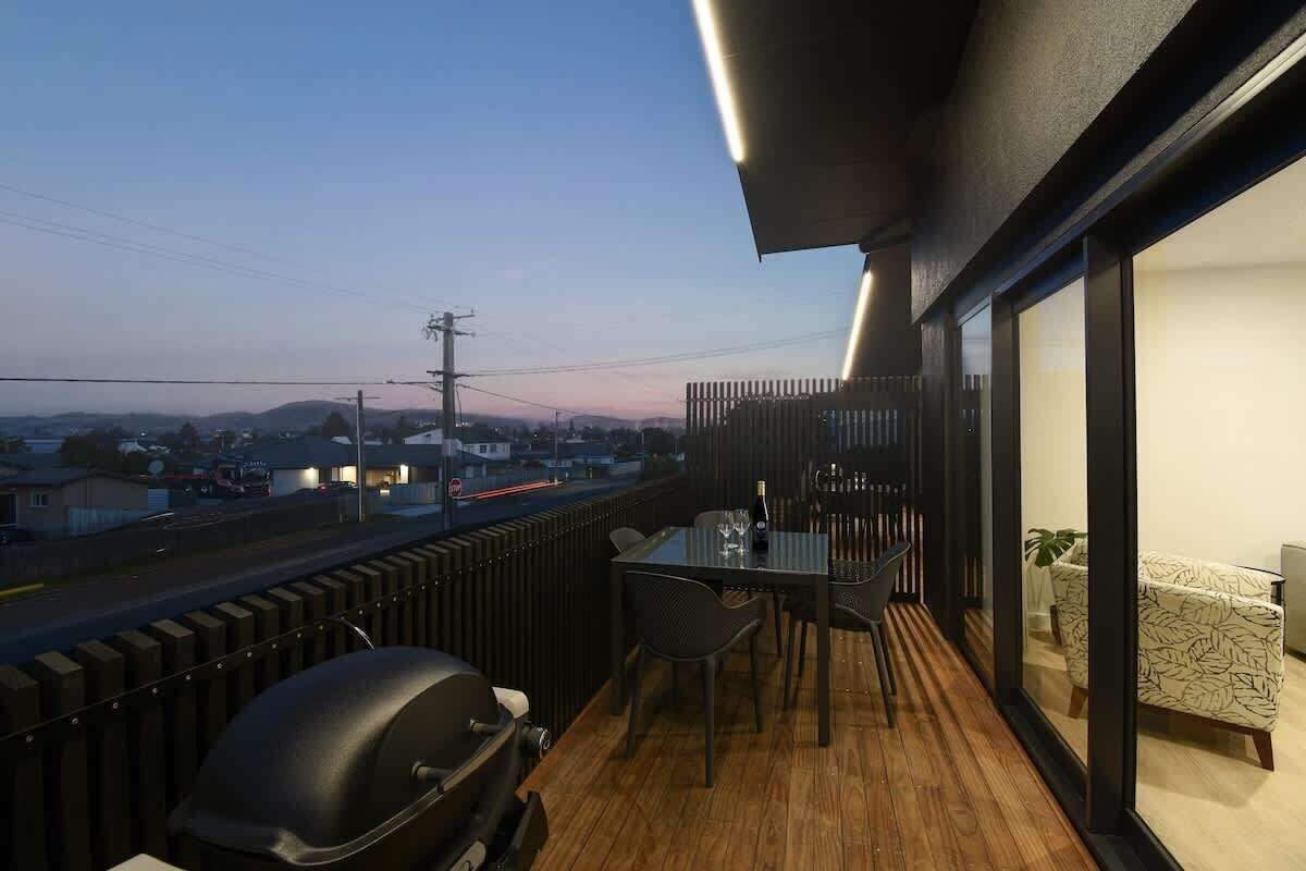 A balcony with a table and chairs, a grill, and a view of the city at dusk.