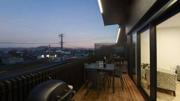 A balcony with a table and chairs, a grill, and a view of the city at dusk.