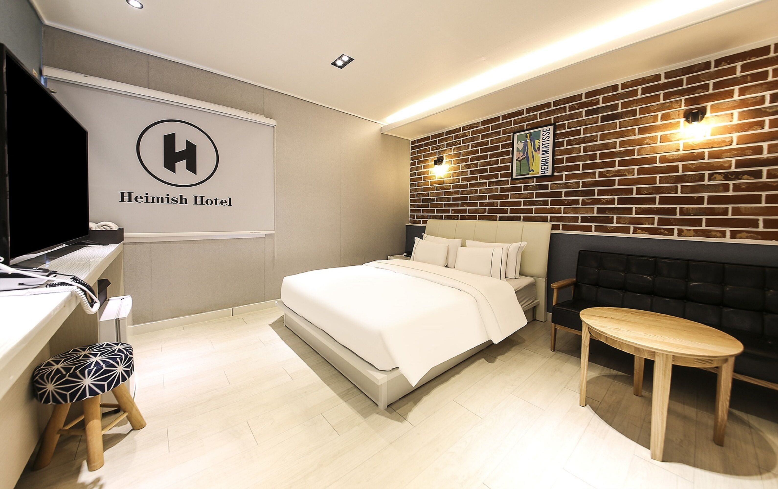 A hotel room with a bed, a desk with a computer, a small stool, a wooden table, and a brick wall with a framed picture.