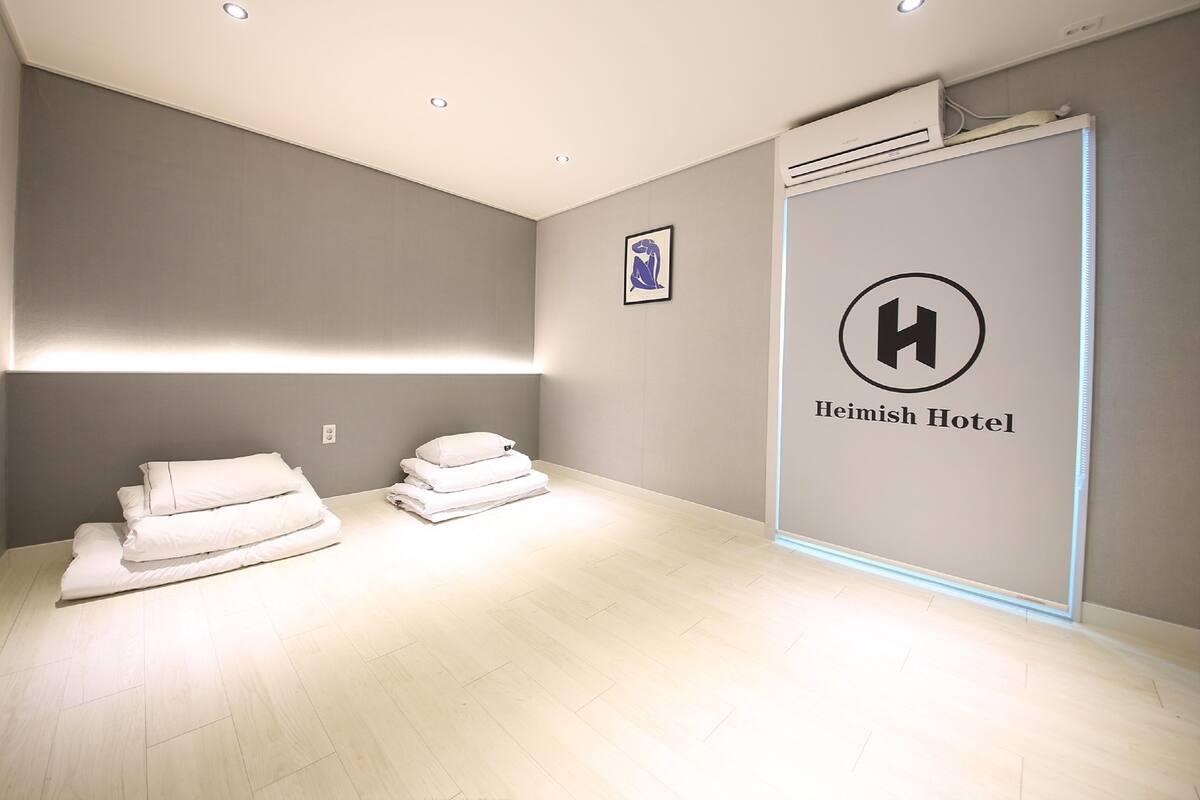 Quarto de hotel com design minimalista, com cama com roupa de cama branca e bem arrumada, unidade de ar condicionado na parede e uma obra de arte emoldurada acima da cama.