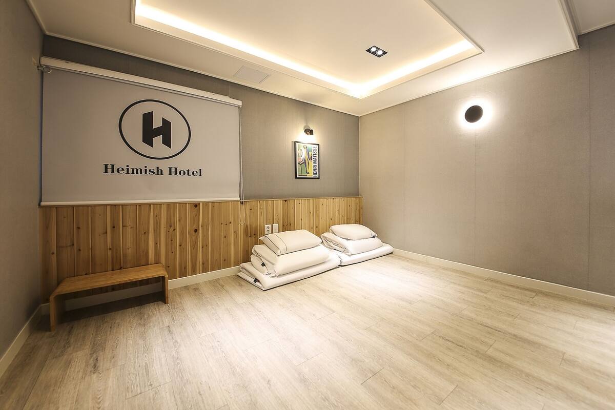 Quarto de hotel com um banco de madeira, uma cama com roupa branca, um cartaz grande com um logotipo e uma luminária fixada na parede.