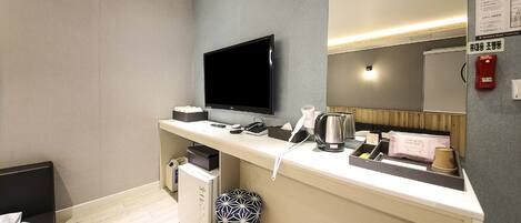 A modern hotel room with a flat-screen TV, a desk with a kettle, a hairdryer, and a small stool.