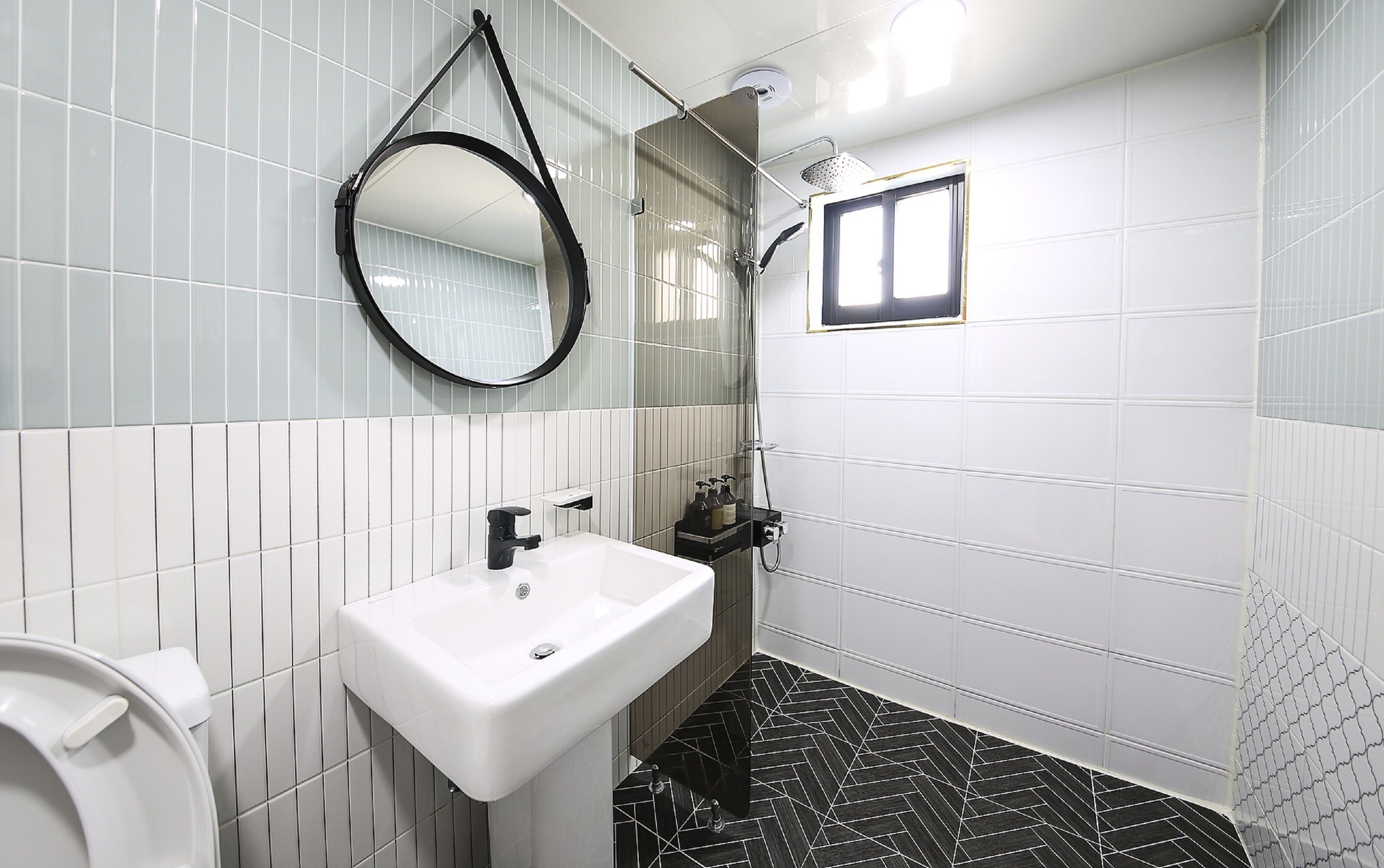 A bathroom with white tiled walls, a black and white patterned floor, a white sink, a toilet, a mirror, and a shower area.