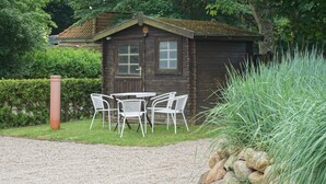 Une cabane en bois rustique, avec un chemin de gravier menant à une table et des chaises en métal peintes en blanc.