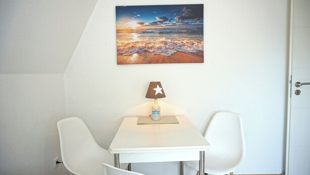 Une pièce avec une table blanche, deux chaises blanches, une lampe fixée au mur et un tableau représentant un coucher de soleil.