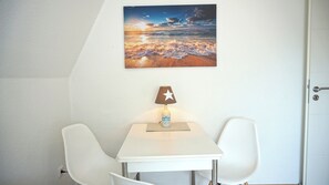 Une pièce avec une table blanche, deux chaises blanches, une lampe fixée au mur et un tableau représentant un coucher de soleil.