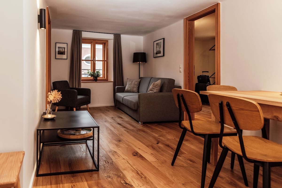 Una sala de estar moderna con un sofá, una mesa de comedor con sillas, una mesita con un florero y una ventana con cortinas.