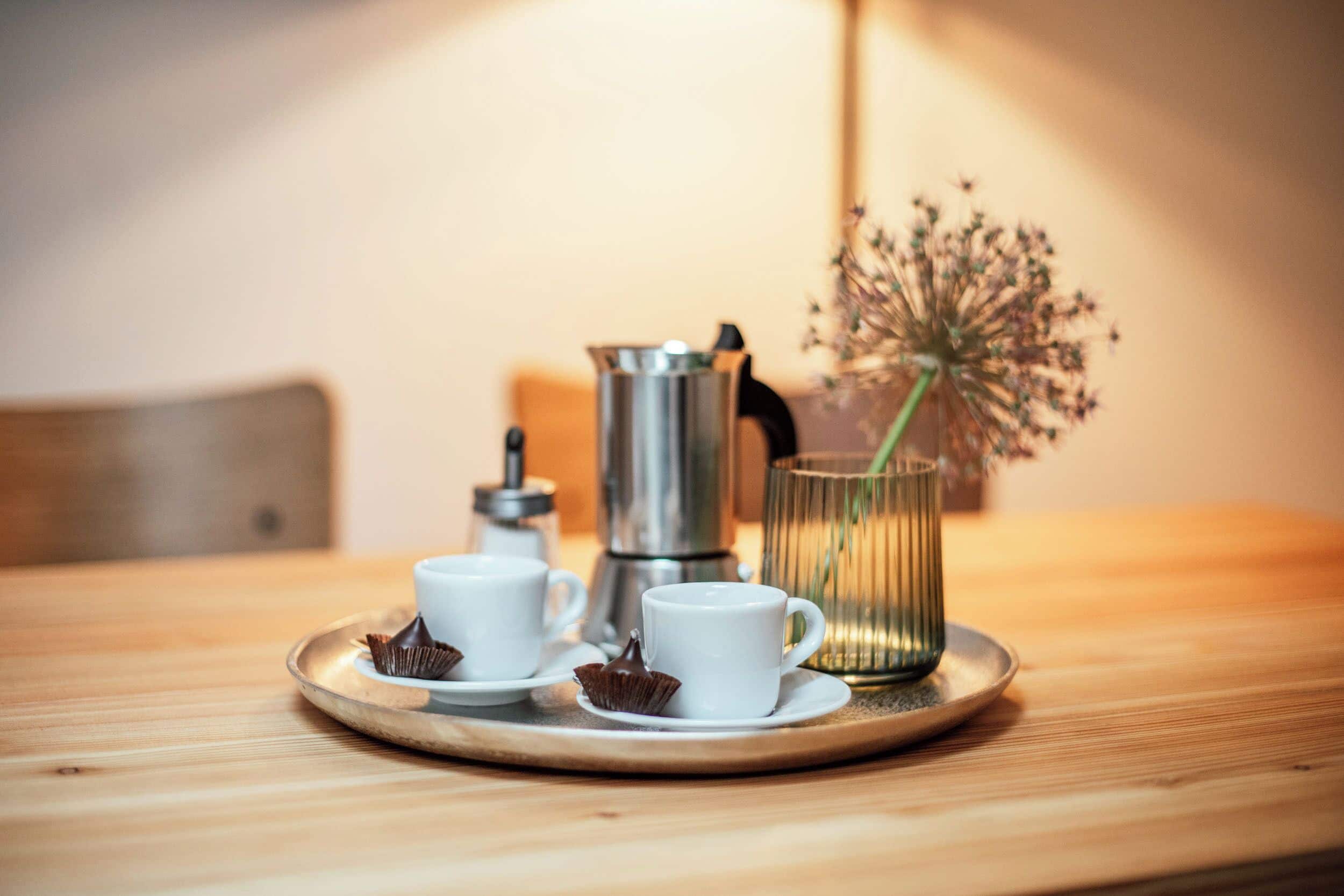 Une table en bois sur laquelle se trouve un plateau avec deux tasses blanches, une cafetière en métal et un vase en verre contenant des fleurs séchées.