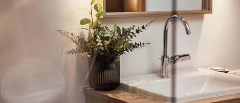 A bathroom with a wooden vanity, a white sink, a modern faucet, and a potted plant.