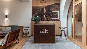 Un intérieur moderne avec un comptoir d’accueil en bois portant l’inscription « The Salt », un canapé gris et une fresque murale représentant un paysage montagneux.