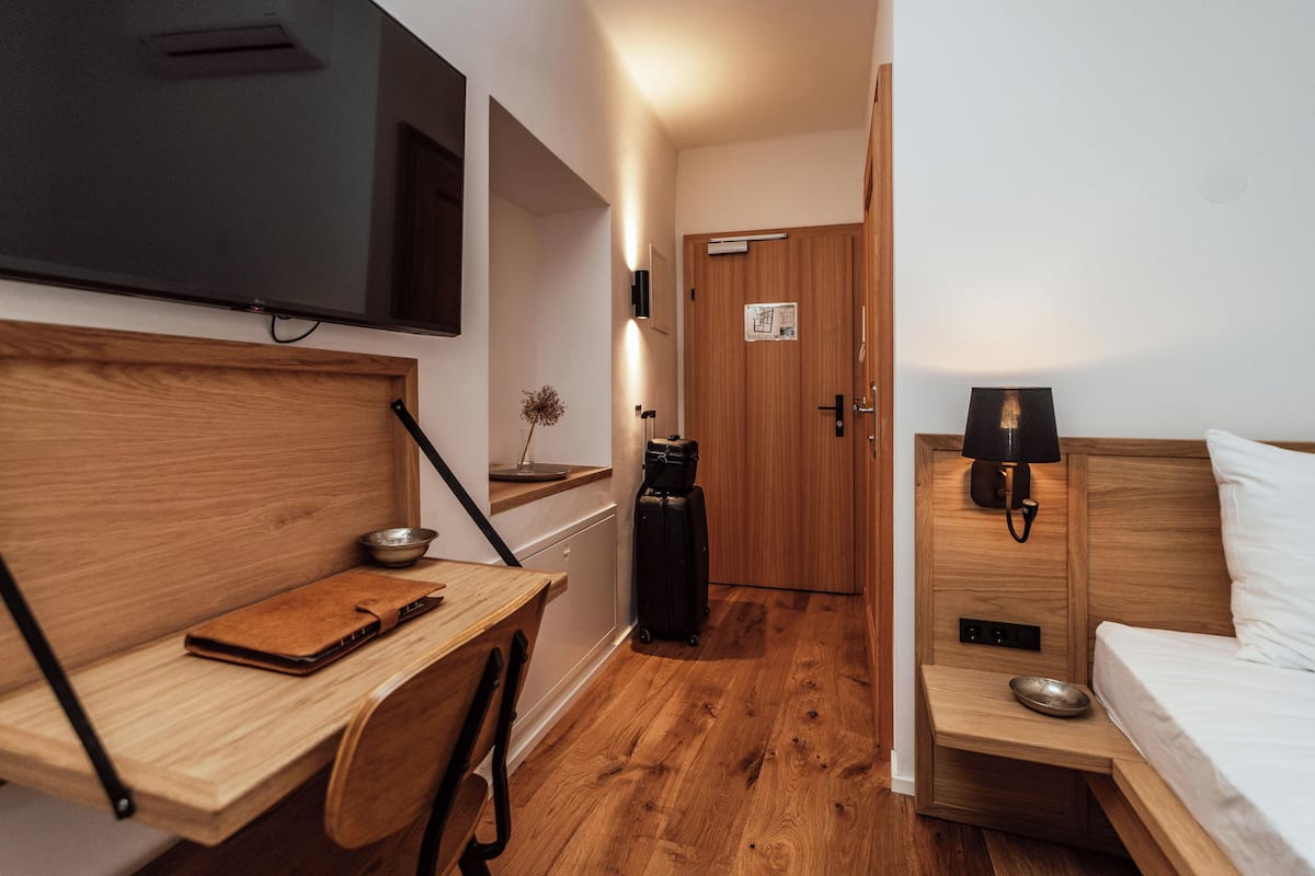 Una habitación de hotel compacta con un escritorio de madera, una cama con mesita de noche, un televisor montado en la pared y una puerta con un cartel.