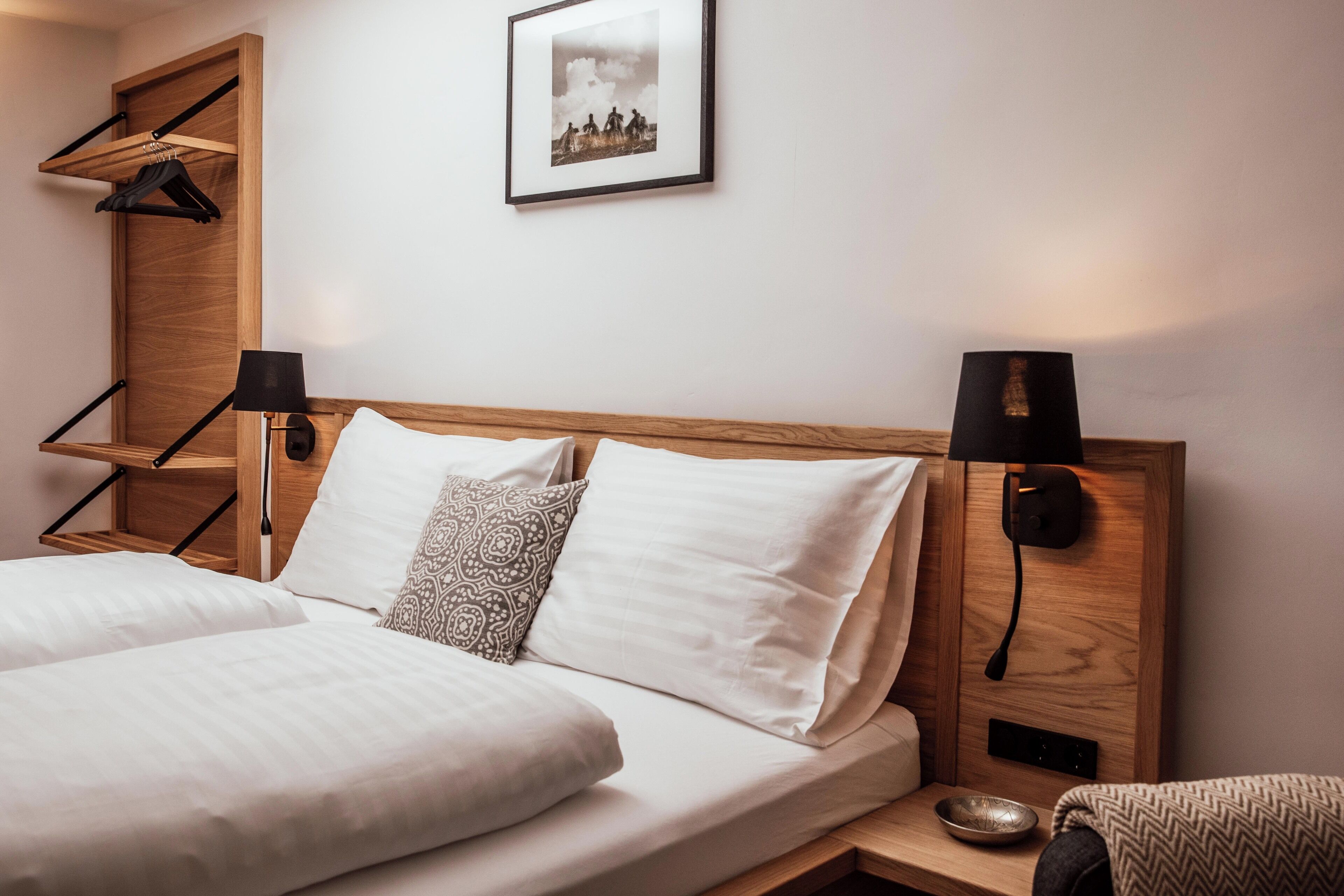 Una habitación de hotel con cabecera de madera, dos mesitas de noche, cama con ropa blanca, almohada decorativa estampada, un cuadro en la pared y una pequeña estantería con un cuenco y un libro.