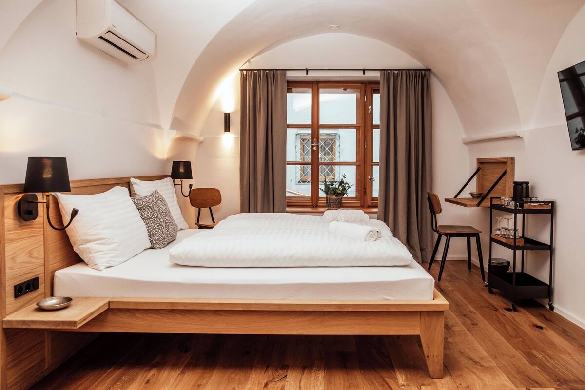 Un dormitorio moderno con una cama de madera, una mesita de noche pequeña, una silla, un escritorio con una silla, una estantería con vasos y una ventana con cortinas.