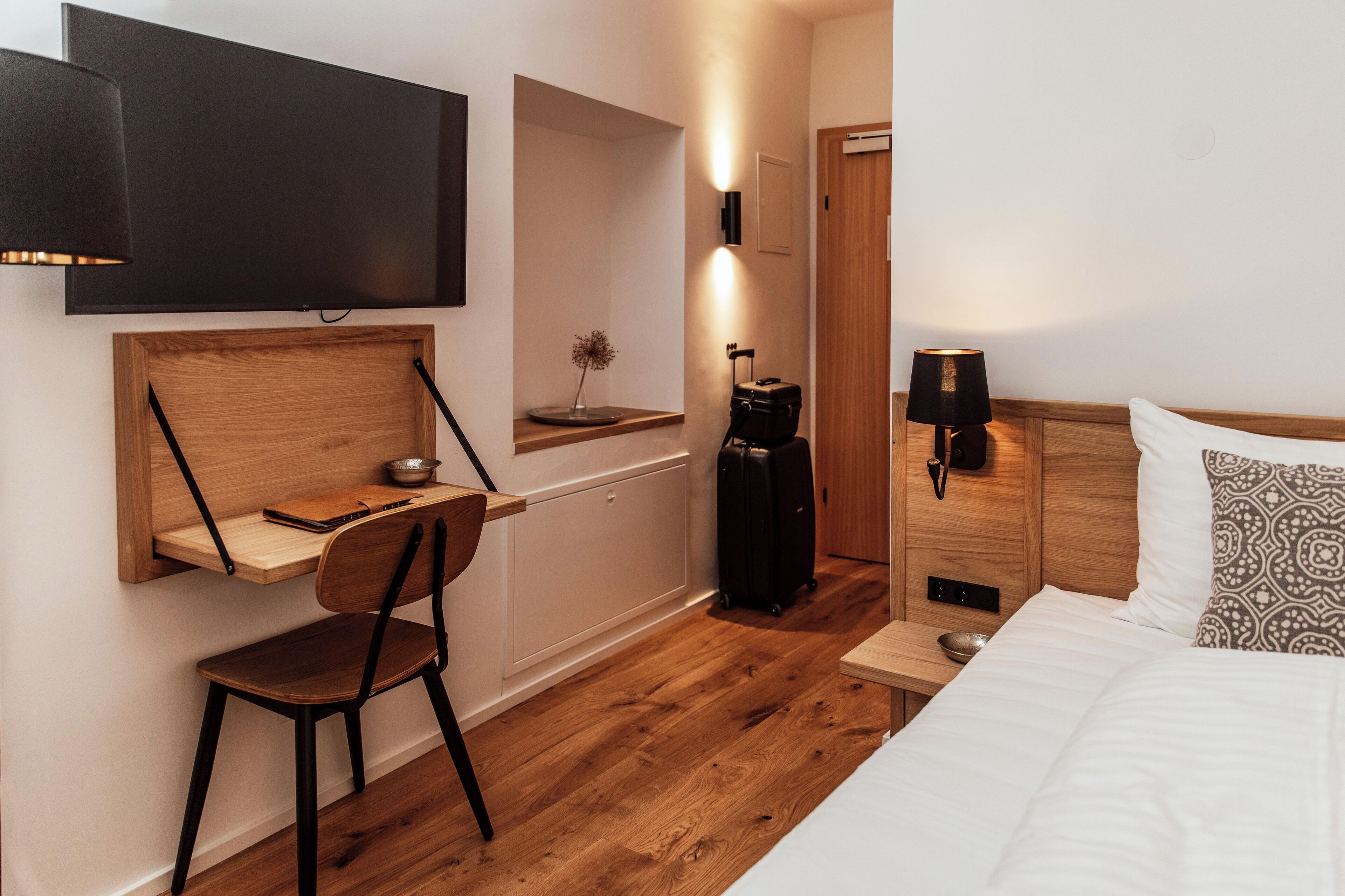 A hotel room with a wooden desk, a chair, a bed with patterned pillows, a TV mounted on the wall, and a suitcase on the floor.