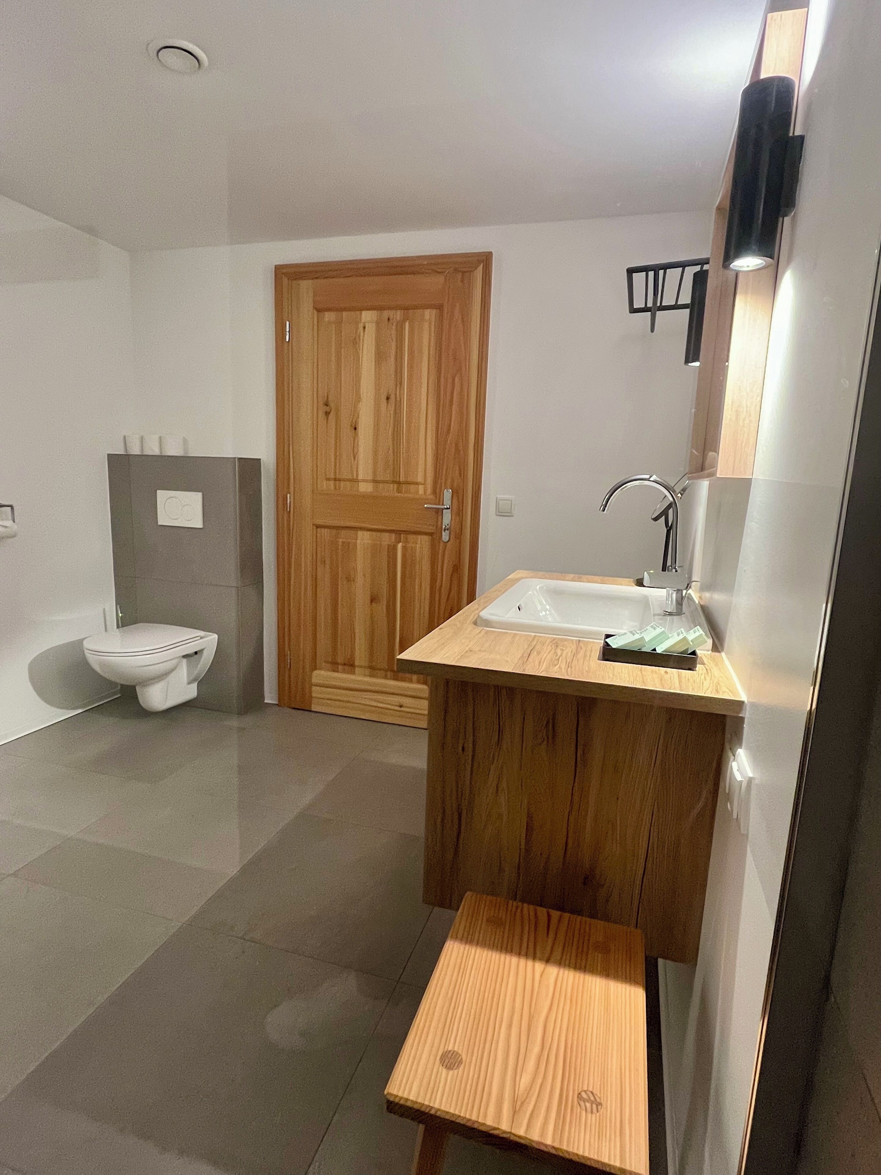 Un baño moderno con puerta de madera, un inodoro, un lavabo con espejo y un pequeño taburete de madera.