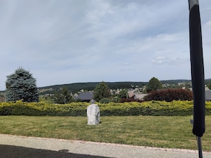 A grassy area with a stone sculpture, a hedge, and a view of houses and hills in the distance.