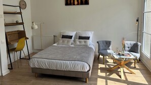A modern bedroom with a large bed, a small round table, a chair, a radiator, a clock, and a window with a view. - La Pléïade Dorée - Hyper Centre, Avec Cours Privative. Animaux Acceptés ! (Amiens)