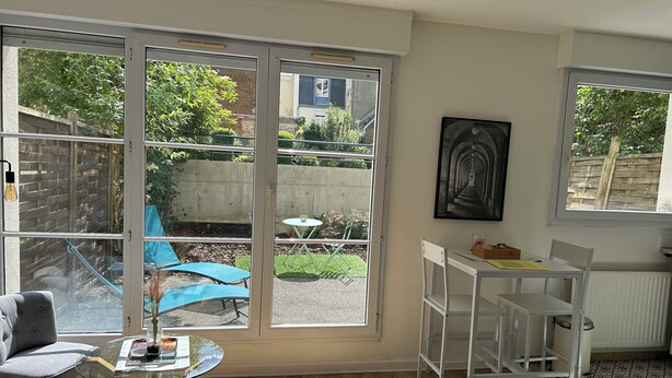 A room with a large sliding glass door leading to a garden, a white chair, a small round table, and a framed picture on the wall. - La Pléïade Dorée - Hyper Centre, Avec Cours Privative. Animaux Acceptés ! (Amiens)