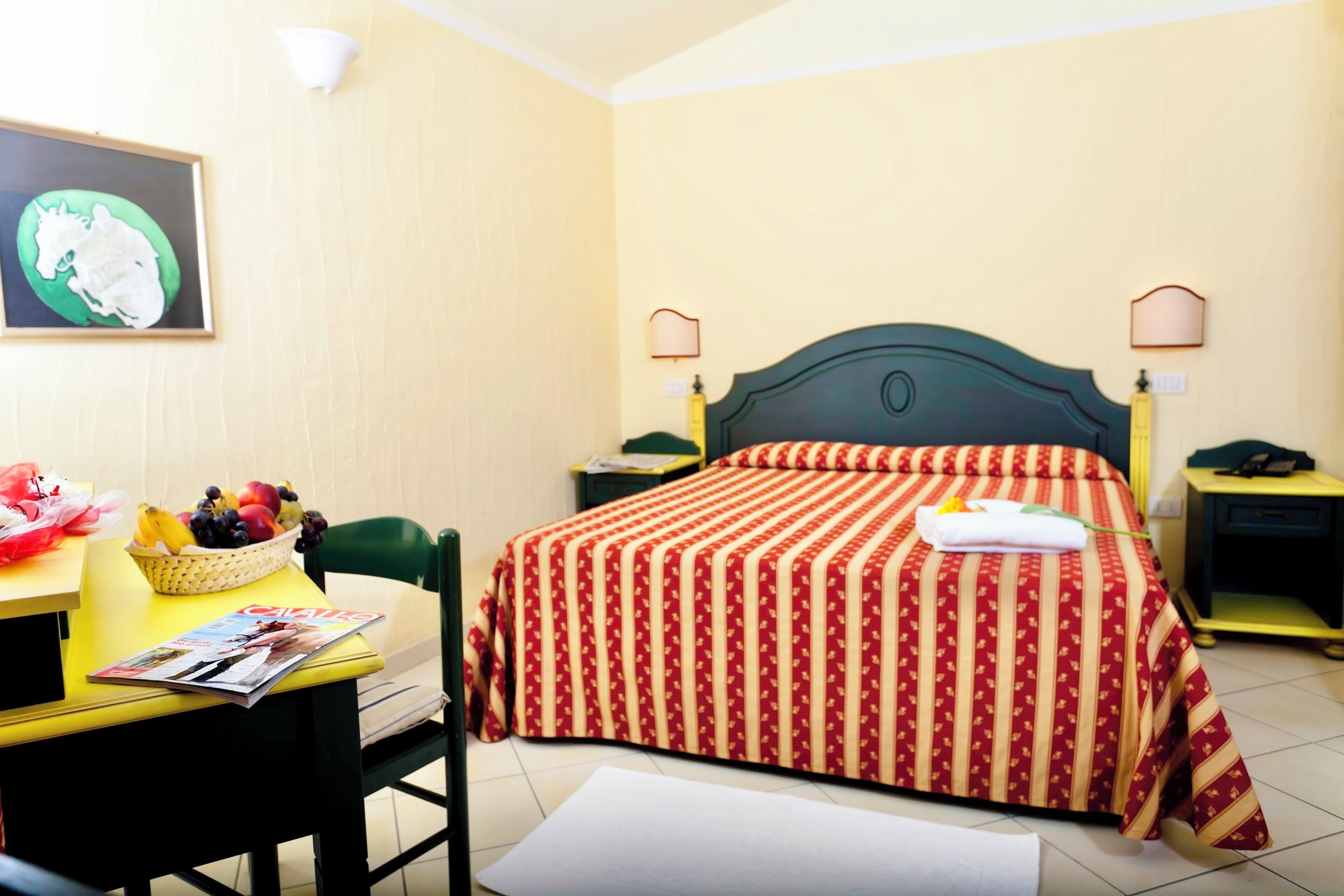 A hotel room with a bed, a desk, a chair, a table with a fruit basket, and a framed picture on the wall.