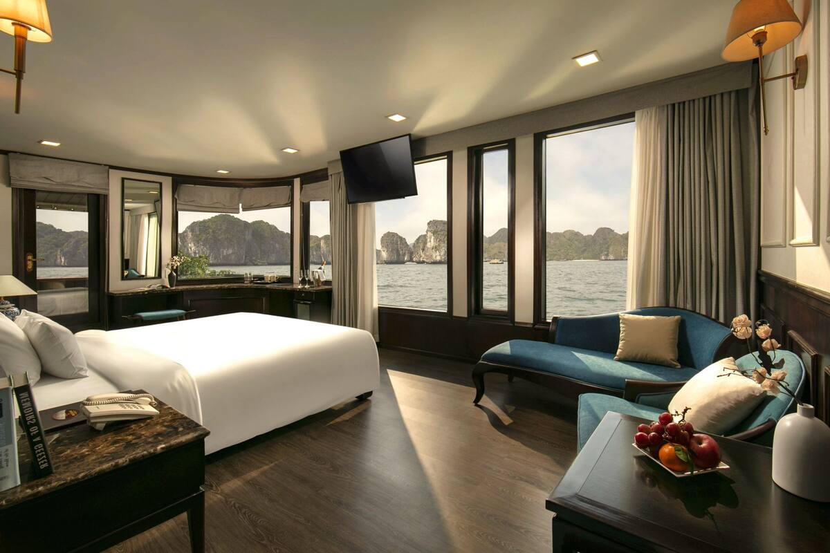 A modern hotel room with a large bed, a seating area with a sofa and armchairs, a desk with a computer, and a TV. The room has large windows offering a view of the sea and cliffs.