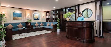 A reception area with a wooden floor, a blue sofa with brown cushions, a reception desk, and a large potted plant.