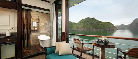 A balcony with a view of a lake and hills, a sofa, a table with a book, and a bathroom with a bathtub.