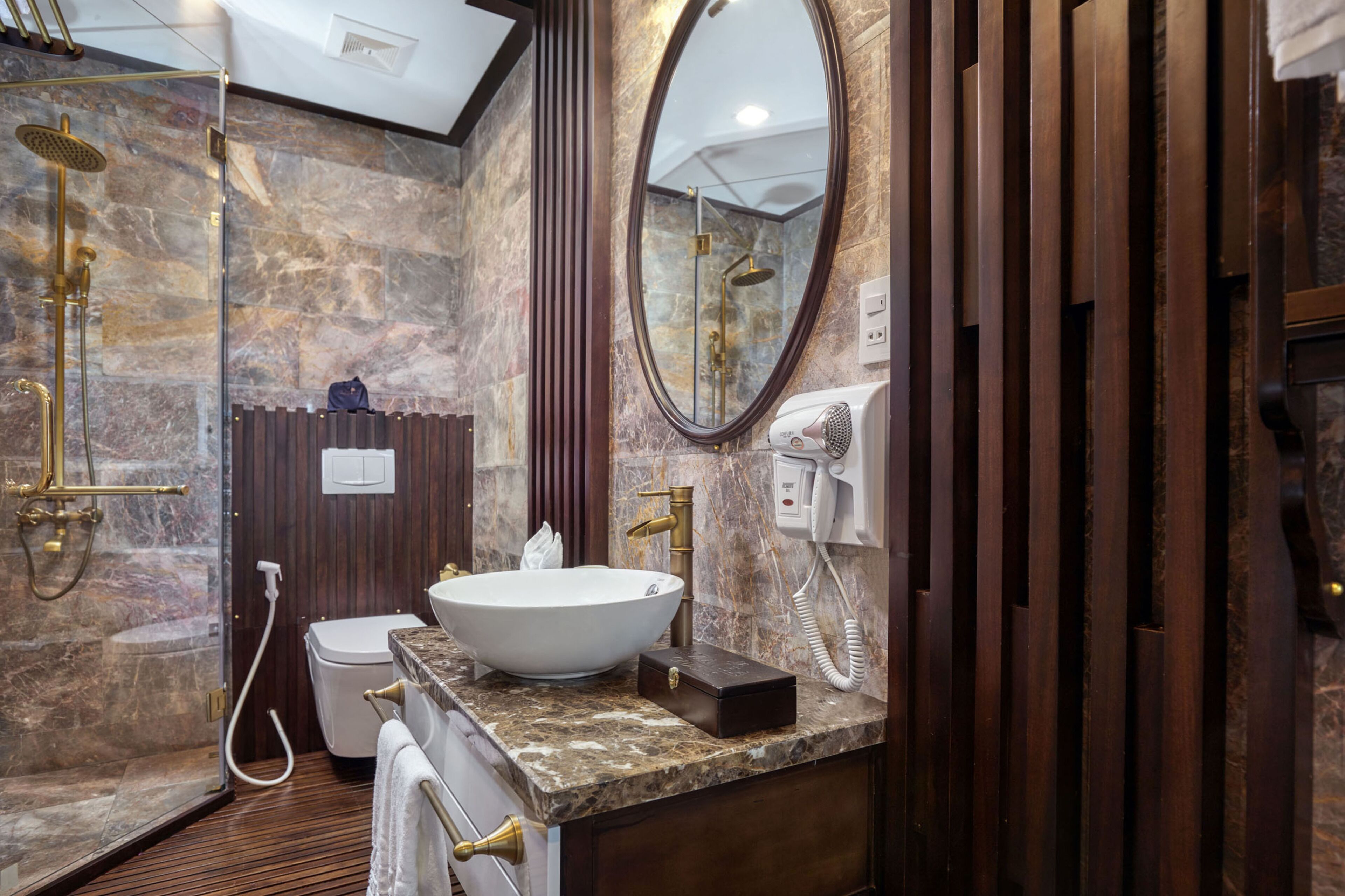 Un baño con pared de mármol, un espejo ovalado grande, un lavabo, un inodoro y una ducha con cabezal de ducha manual.