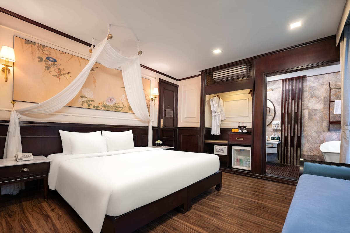 A hotel room with a large bed, a bedside table with a lamp, a minibar, a bathroom with a mirror, and a large painting on the wall.