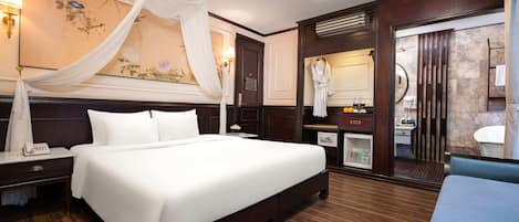 A hotel room with a large bed, a bedside table with a lamp, a minibar, a bathroom with a mirror, and a large painting on the wall.