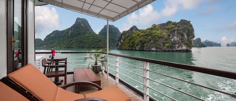 A balcony with lounge chairs and a table, overlooking a body of water and green mountains.