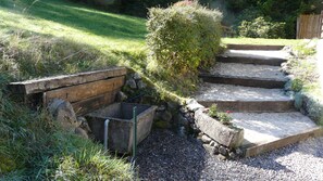 Un jardín con un sendero de grava, escalones de madera y una jardinera con flores rojas.