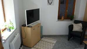 Ein Wohnzimmer mit einem Flachbildfernseher auf einem Holzständer, einem schwarzen Sessel mit Kissen, einer Topfpflanze und einem Fenster mit einem dekorativen Kranz.