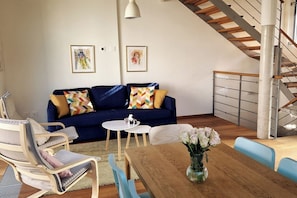 Un salon avec un canapé bleu, une table à manger, un vase de fleurs, un escalier et deux œuvres d’art encadrées accrochées au mur.