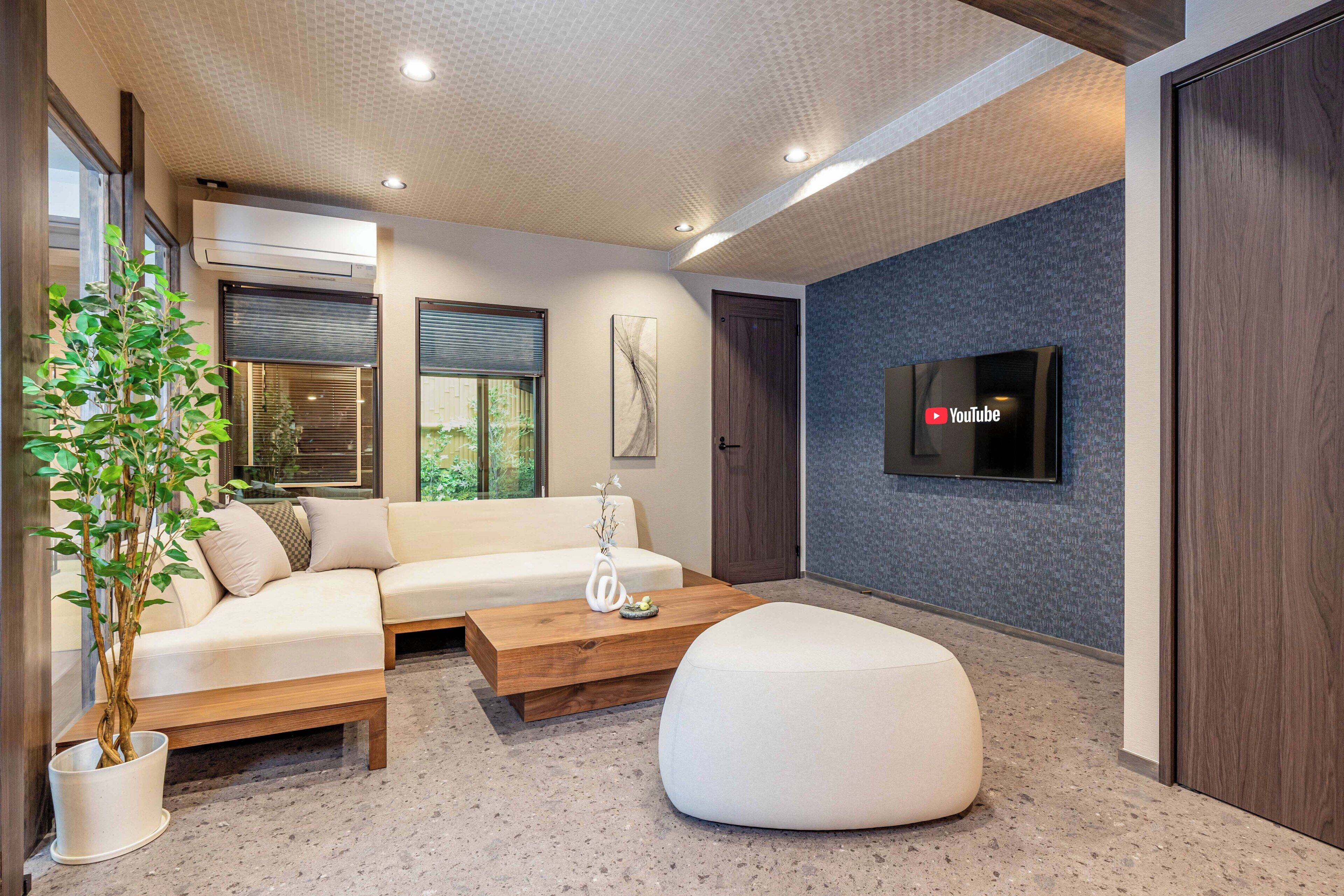 A modern living room with a sofa, coffee table, and ottoman, a flat-screen TV mounted on the wall, and a potted plant.