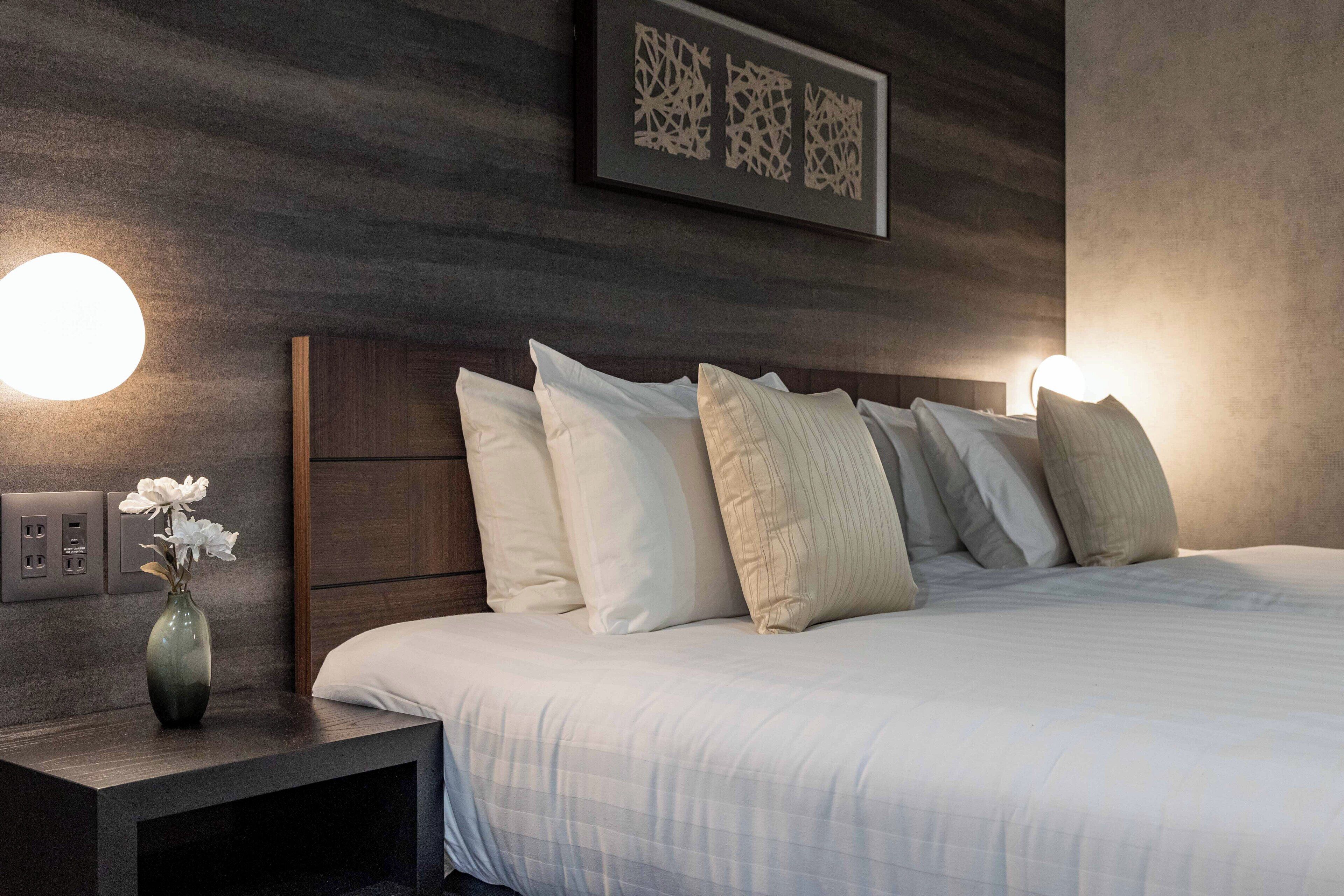 A modern hotel room with a large bed, a nightstand with a vase of flowers, and wall-mounted lighting.