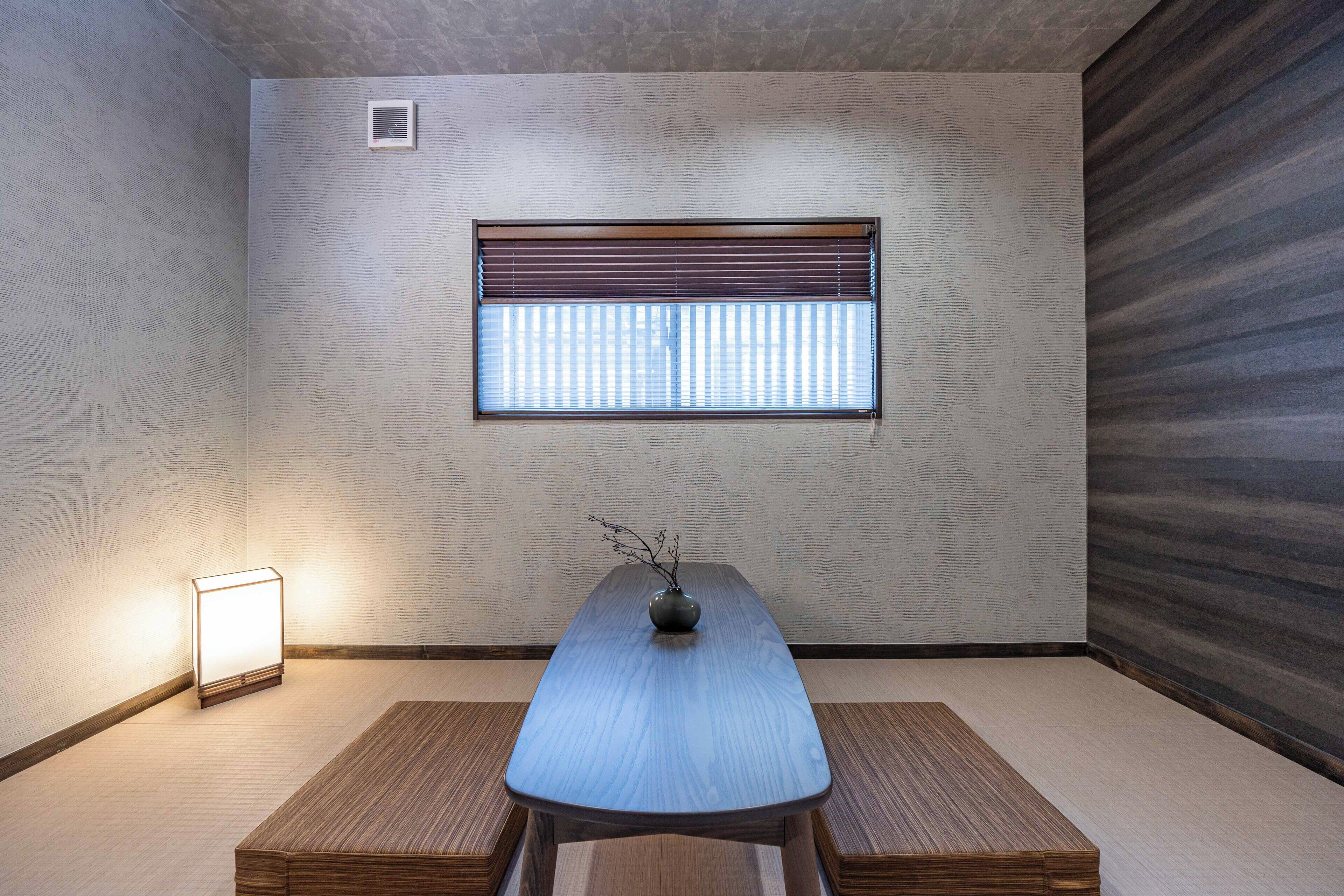 A minimalist room with a wooden table, two benches, a vase with branches, a window with blinds, and a wall-mounted light.