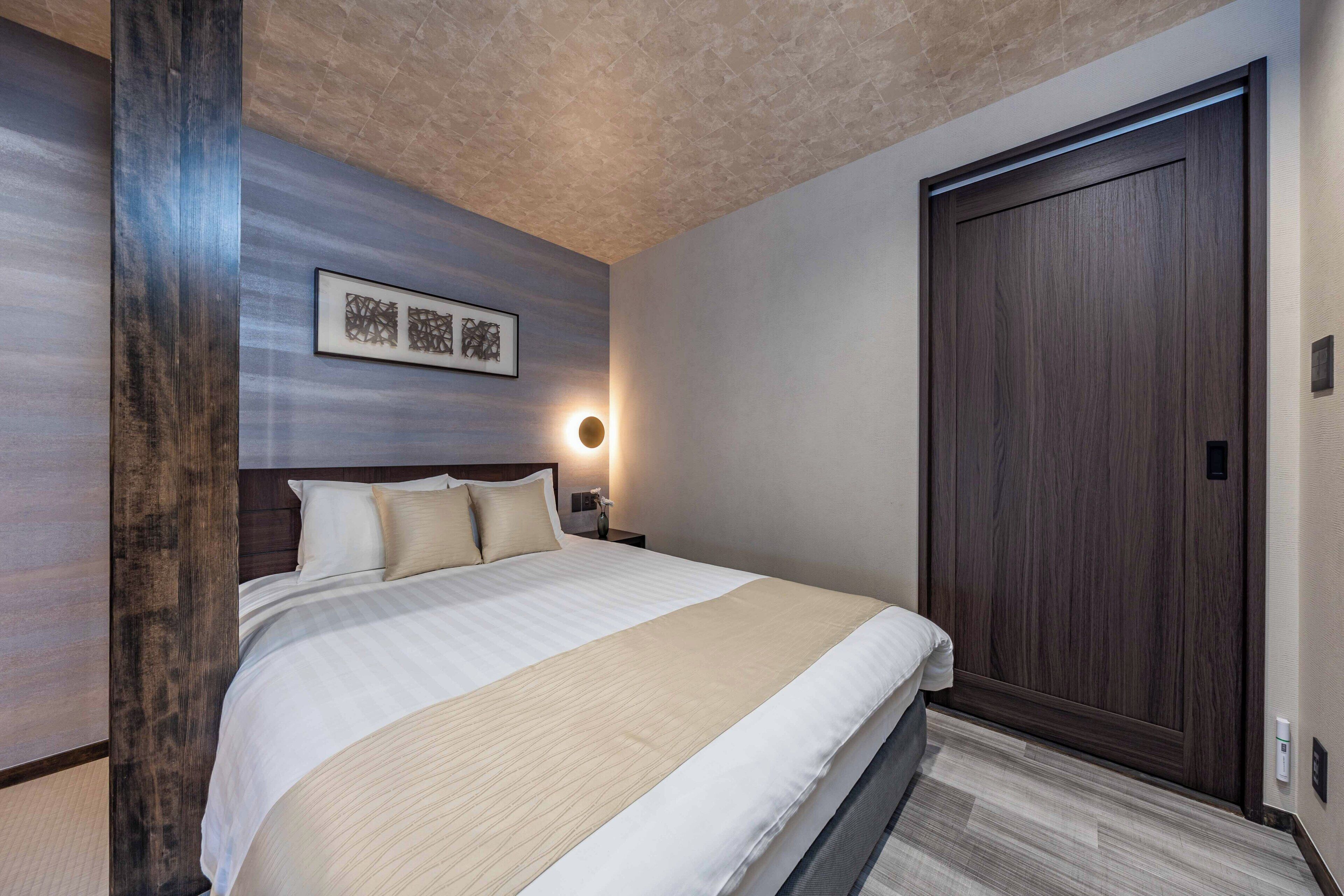 A hotel room with a large bed, a wooden headboard, a framed artwork on the wall, a wall-mounted light, and a dark wooden door.