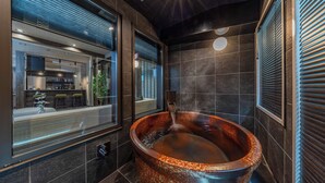 A modern bathroom with a large, rustic bathtub, a visible kitchen area through a glass partition, and a contemporary design with dark tiles and a sleek ceiling. - JAPAVISTA Hisho (Osaka)